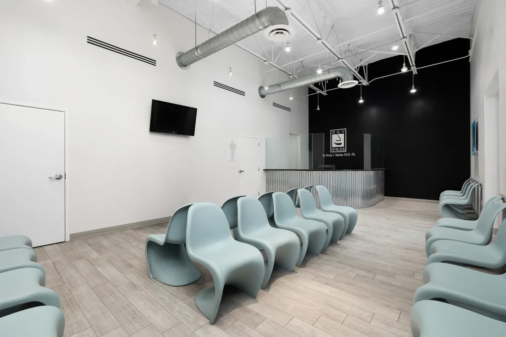 The image shows an interior space with rows of blue chairs facing forward, arranged inside a waiting area with a tiled floor, and there s a television screen mounted on one wall.