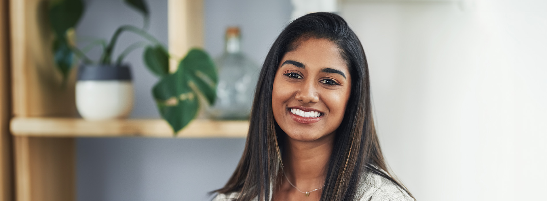 The image features a smiling woman with dark hair, wearing a white top, standing indoors with a plant in the background.