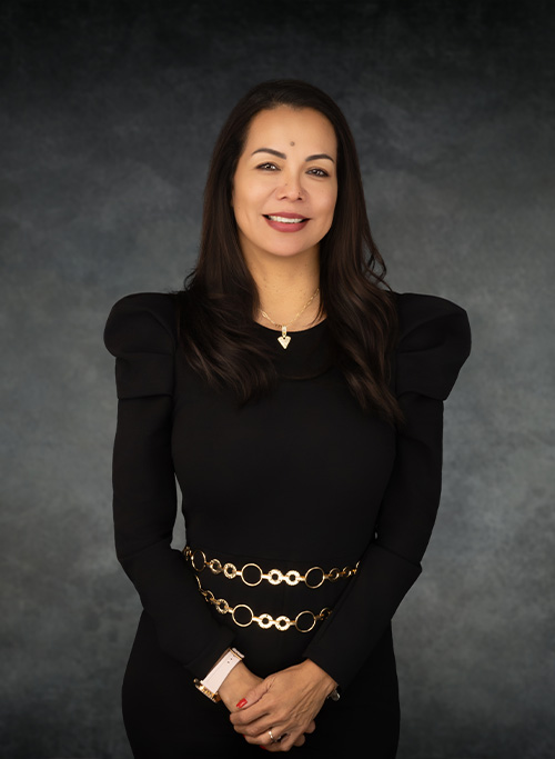The image shows a woman standing against a dark background with her hands clasped together at her waist, wearing a black outfit and accessories, and she has a neutral expression on her face.