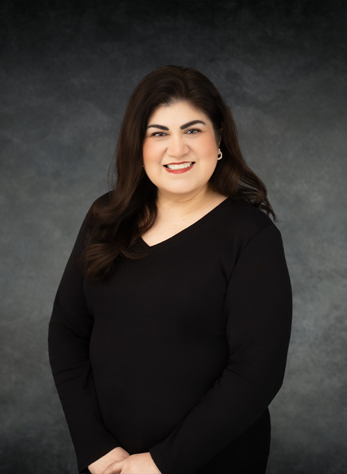 A woman with dark hair wearing a black dress is standing against a grey background.