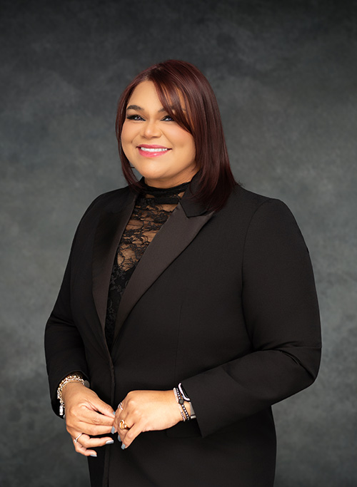 The image features a woman dressed in formal attire, standing against a dark background with a neutral expression on her face.