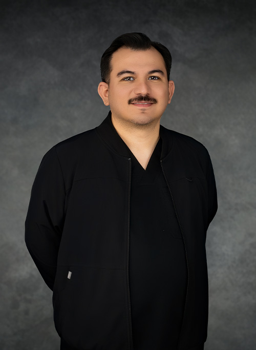The image shows a man standing against a background with a formal pose. He has dark hair, a mustache, and is wearing a black jacket with an open collar.