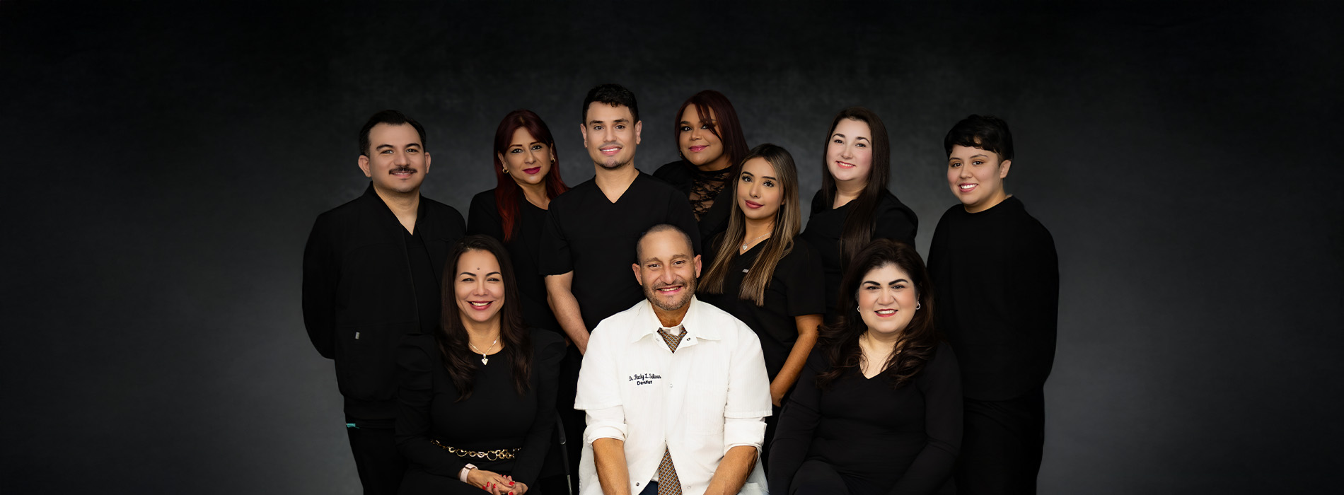 A group of people posing together in front of a dark background, with one individual standing out in the center wearing a white shirt and holding a microphone.